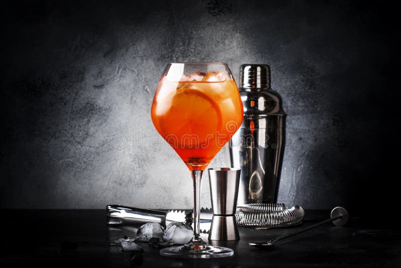 Aperol spritz cocktail in big wine glass with orange and ice, summer alcoholic cold drink, dark bar counter with steel bar tools