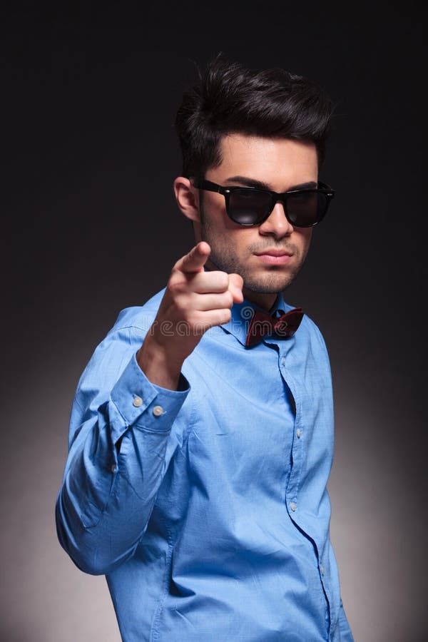 Serious looking young male pointing at the camera and standing while wearing sunglasses and suit on grey studio background. Serious looking young male pointing at the camera and standing while wearing sunglasses and suit on grey studio background