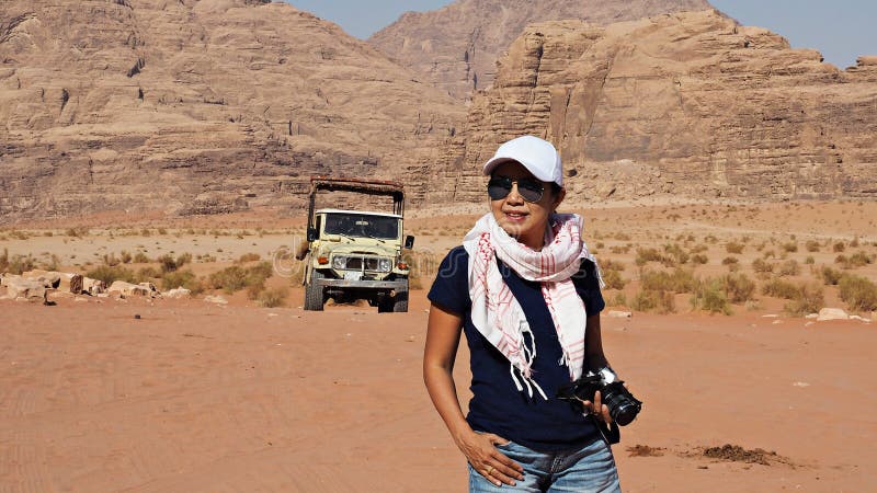 Appareil-photo de transport de femme asiatique de touristes en désert de Wadi Rum, Jordanie avec la voiture de safari derrière image stock Appareil-photo de transport de femme asiatique de touristes en désert de Wadi Rum, Jordanie avec la voiture de safari derrière image stock