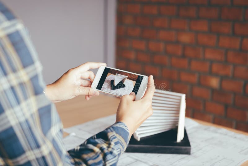 Architect man holding smartphone taking a photo of an architecture model sent to client and working in home office. stock photo Architect man holding smartphone taking a photo of an architecture model sent to client and working in home office. stock photo