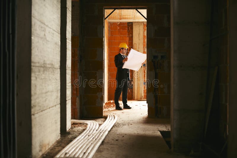 Engineer at work in construction site, standing in apartment building and looking at blueprints and plans. Engineer at work in construction site, standing in apartment building and looking at blueprints and plans