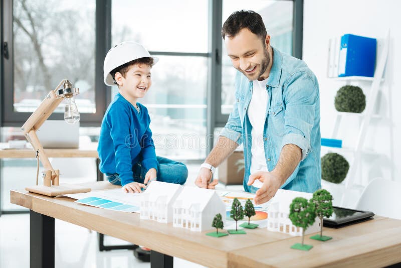 Clear explanation. Cheerful young architect explaining the basics of his work to his little son sitting on the top of the table and listening to him with a fond smile. Clear explanation. Cheerful young architect explaining the basics of his work to his little son sitting on the top of the table and listening to him with a fond smile