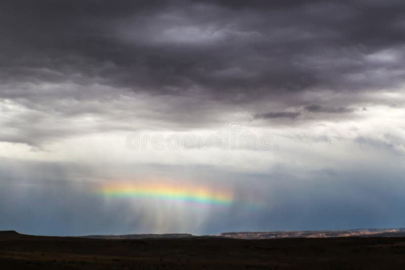 Arcobaleno orizzontale attraverso il mezzo di un groppo della pioggia vicino all'orizzonte in deserto con le scogliere distanti a fotografia stock Arcobaleno orizzontale attraverso il mezzo di un groppo della pioggia vicino all'orizzonte in deserto con le scogliere distanti a fotografia stock