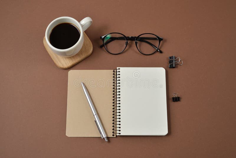 Area di lavoro del modello con gli articoli per ufficio, la penna, la tazza di caffè, il blocco note ed i vetri su fondo marrone immagine stock Area di lavoro del modello con gli articoli per ufficio, la penna, la tazza di caffè, il blocco note ed i vetri su fondo marrone immagine stock