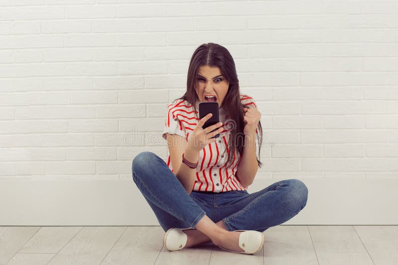 Angry young woman screaming on mobile phone, sitting cross-legged, Isolated white brick wall background.. Negative human emotions, facial expressions, life perception. Angry young woman screaming on mobile phone, sitting cross-legged, Isolated white brick wall background.. Negative human emotions, facial expressions, life perception
