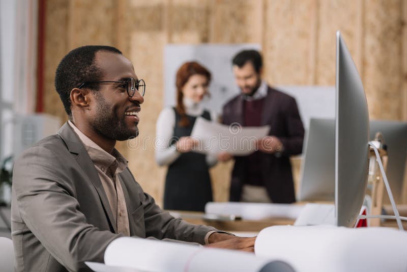 arquitecto afroamericano feliz que trabaja con el ordenador en la oficina moderna mientras que sus colegas imagenes de archivo arquitecto afroamericano feliz que trabaja con el ordenador en la oficina moderna mientras que sus colegas imagenes de archivo