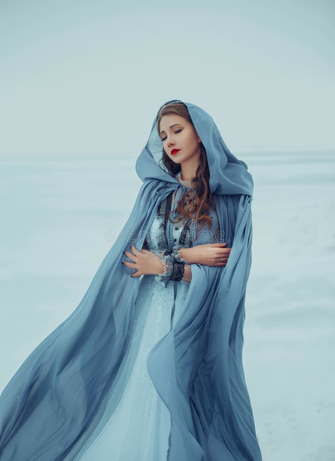 Art photo. Fantasy young woman fairy elf in blue cape with hood stands in cold wind. Winter nature background, white