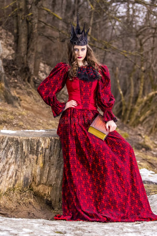 Art Photography. Mysterious Medieval Queen in Red Dress and Spiky Black Crown Posing With Ancient Book in Forest in Early Spring royalty free stock photo Art Photography. Mysterious Medieval Queen in Red Dress and Spiky Black Crown Posing With Ancient Book in Forest in Early Spring royalty free stock photo