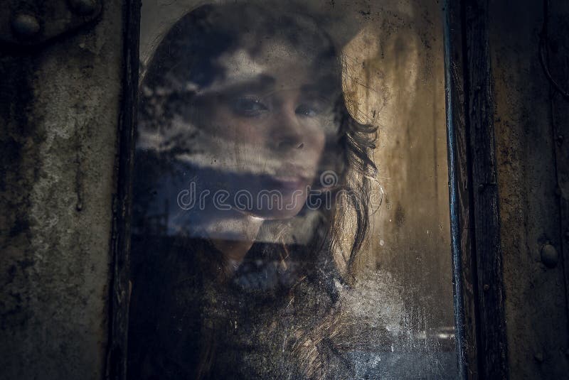 Art portrait of a beautiful young spooky woman, looks through grunge styled rainy window.