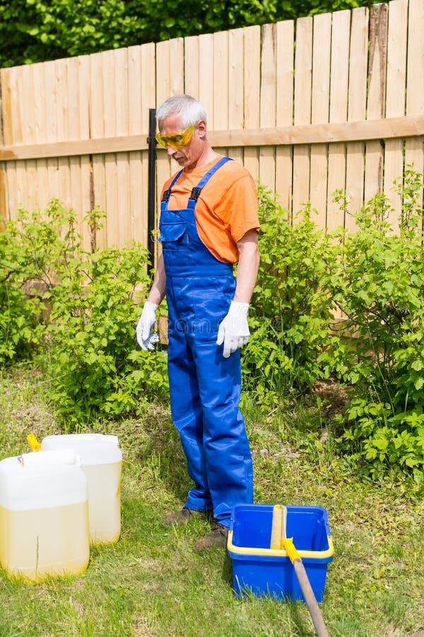 Artesano en el uniforme azul y anaranjado que mira las latas y el cubo de la pintura en el jardín foto de archivo libre de regalías Artesano en el uniforme azul y anaranjado que mira las latas y el cubo de la pintura en el jardín foto de archivo libre de regalías