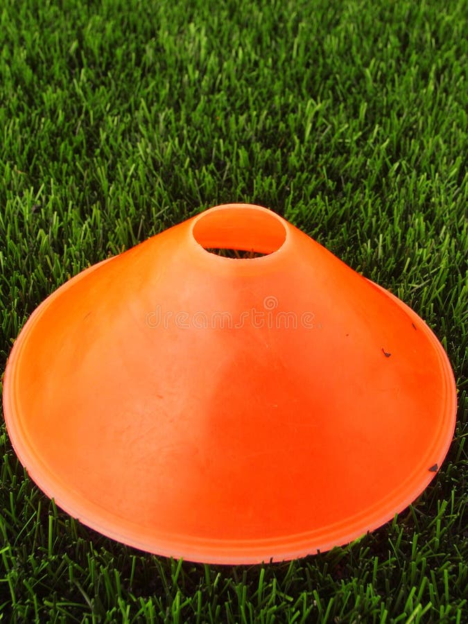 Artificial green plastic grass in background with bright orange plastic cone. Mark on winter footbal playground.