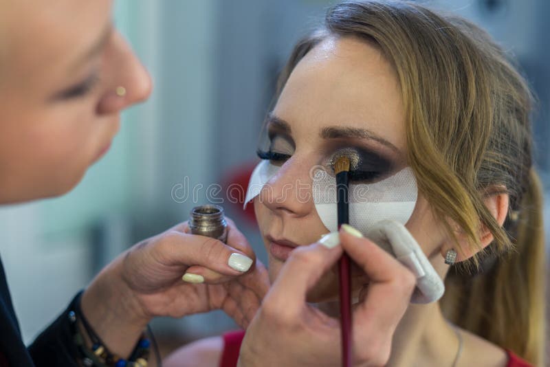 Artista de maquillaje que hace maquillaje ahumado de los ojos a la chica joven hermosa foto de archivo Artista de maquillaje que hace maquillaje ahumado de los ojos a la chica joven hermosa foto de archivo