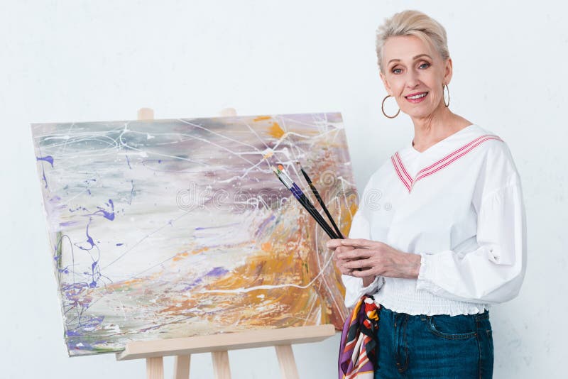 artista femminile elegante senior che tiene i pennelli e che sta vicino alla pittura sul cavalletto fotografia stock libera da diritti artista femminile elegante senior che tiene i pennelli e che sta vicino alla pittura sul cavalletto fotografia stock libera da diritti