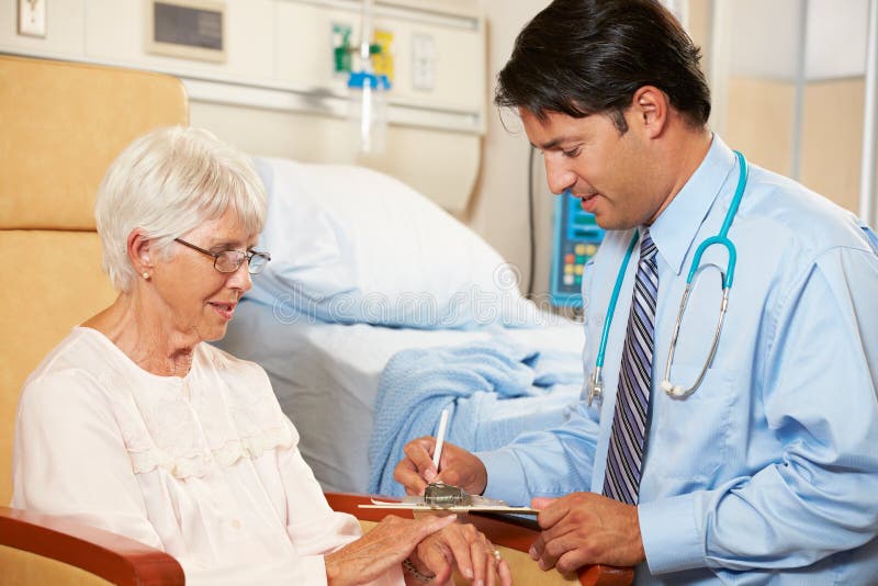 Doctor Taking Notes From Senior Female Patient Seated In Chair By Hospital Bed. Doctor Taking Notes From Senior Female Patient Seated In Chair By Hospital Bed