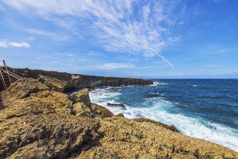 Rocky coastline with waves crashing against shore in Arikok National Park under blue sky with white clouds. Aruba.