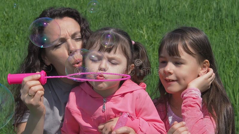 As crianças com a mamã na grama verde estão fundindo bolhas de sabão Família em um parque do verão Mamã e filhas que jogam na nat