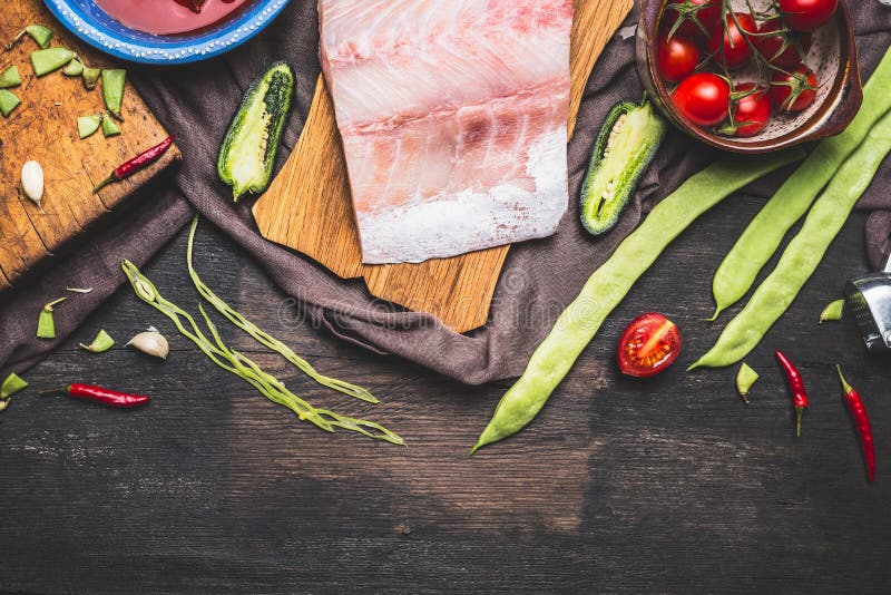 As faixas de peixes tornam côncava o cozimento e a preparação com feijões verdes, tomates e ingredientes no fundo de madeira escu imagens de stock As faixas de peixes tornam côncava o cozimento e a preparação com feijões verdes, tomates e ingredientes no fundo de madeira escu imagens de stock