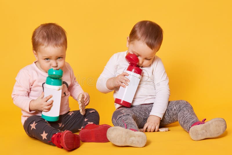 As irmãs gêmeas cansadas sentam-se no assoalho ao comer e ao beber As crianças brincalhão bonitos são furadas no meio do dia pouc imagem de stock As irmãs gêmeas cansadas sentam-se no assoalho ao comer e ao beber As crianças brincalhão bonitos são furadas no meio do dia pouc imagem de stock