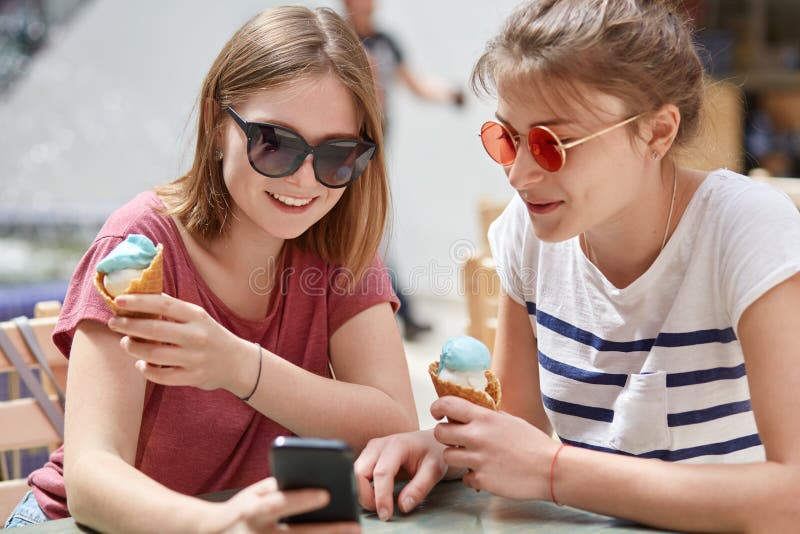 As jovens mulheres bonitas com os olhares felizes, focalizados no telefone esperto, olham o filme em linha, comem o gelado, veste imagens de stock As jovens mulheres bonitas com os olhares felizes, focalizados no telefone esperto, olham o filme em linha, comem o gelado, veste imagens de stock