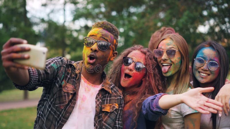 As meninas entusiasmado e os indivíduos dos amigos estão tomando o selfie com caras coloridas e o cabelo usando o smartphone, hom