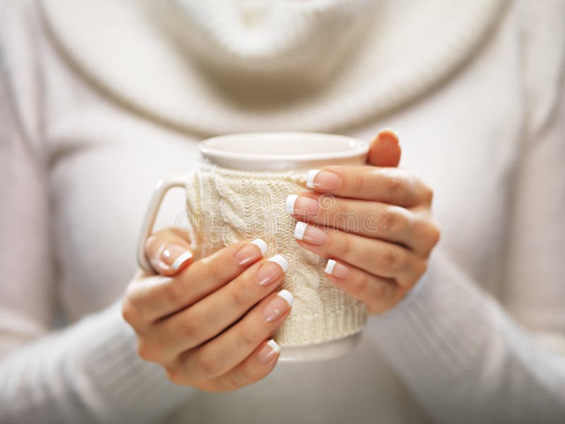 Woman holds a winter cup close up on light background. Woman hands with elegant french manicure nails design holding a cozy knitted mug. Winter and Christmas time concept. Woman holds a winter cup close up on light background. Woman hands with elegant french manicure nails design holding a cozy knitted mug. Winter and Christmas time concept.