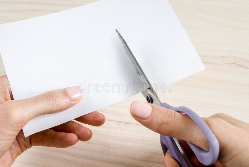 Female hands cutting blank paper or card with a pair of purple handed scissors with wood background. Female hands cutting blank paper or card with a pair of purple handed scissors with wood background