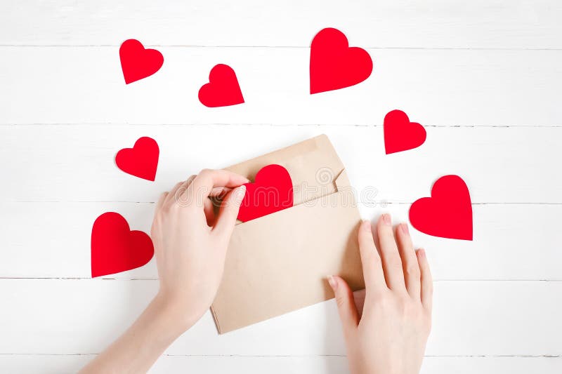 As mãos das mulheres colocam corações vermelhos em um envelope de parabéns sobre um fundo branco de madeira feliz namorados dia fu