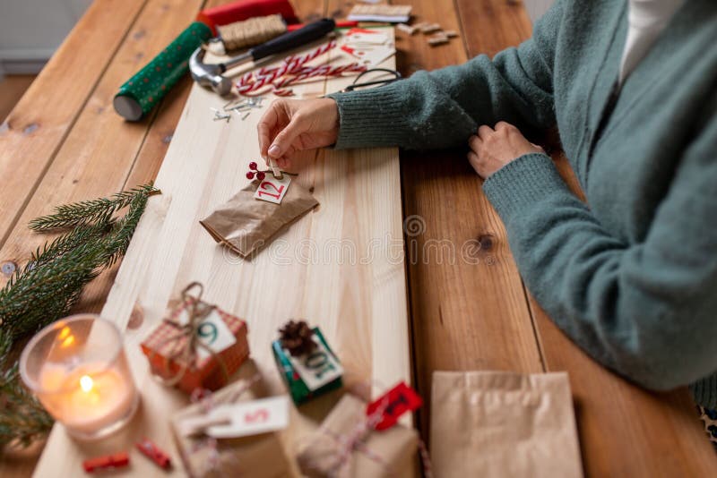 winter holidays and hobby concept - hands with clothespin, craft paper bag and tag making advent calendar on christmas at home. winter holidays and hobby concept - hands with clothespin, craft paper bag and tag making advent calendar on christmas at home