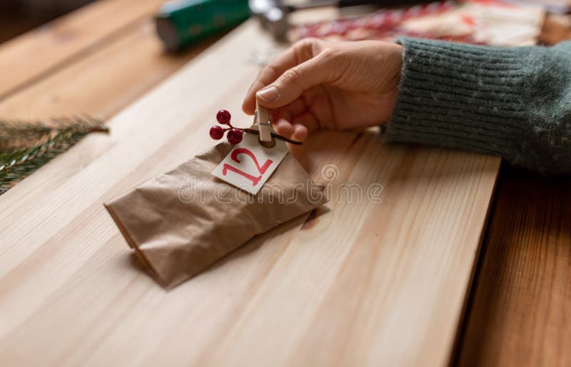 winter holidays and hobby concept - hands with clothespin, craft paper bag and tag making advent calendar on christmas at home. winter holidays and hobby concept - hands with clothespin, craft paper bag and tag making advent calendar on christmas at home