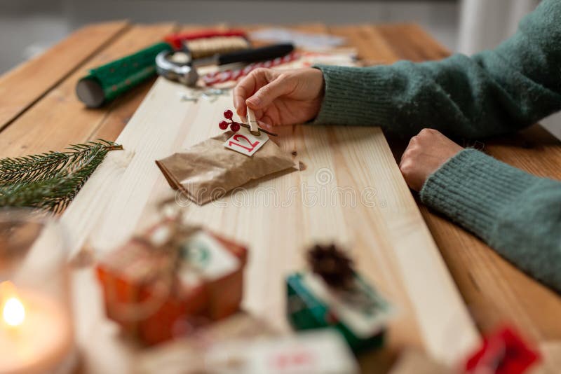 winter holidays and hobby concept - hands with clothespin, craft paper bag and tag making advent calendar on christmas at home. winter holidays and hobby concept - hands with clothespin, craft paper bag and tag making advent calendar on christmas at home