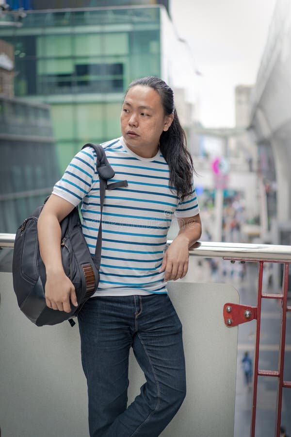 Asain / Thai-Chinese long black hair guy with blue line t-shirt is thinking and holding backpack on his sholder near the sky train stock images Asain / Thai-Chinese long black hair guy with blue line t-shirt is thinking and holding backpack on his sholder near the sky train stock images