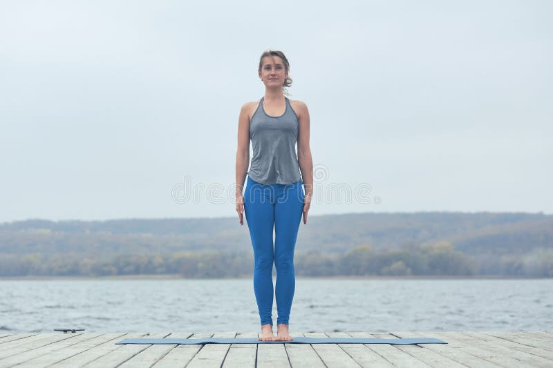 Asana hermoso Tadasana - actitud de la yoga de las prácticas de la mujer joven de la montaña en la cubierta de madera cerca del l fotos de archivo libres de regalías Asana hermoso Tadasana - actitud de la yoga de las prácticas de la mujer joven de la montaña en la cubierta de madera cerca del l fotos de archivo libres de regalías