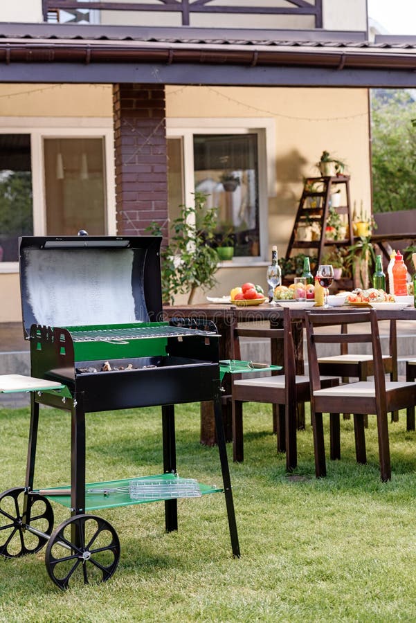 Ase a la parrilla con los carbones que se colocan en césped verde y la tabla servida con los platos y las bebidas detrás fotografía de archivo Ase a la parrilla con los carbones que se colocan en césped verde y la tabla servida con los platos y las bebidas detrás fotografía de archivo