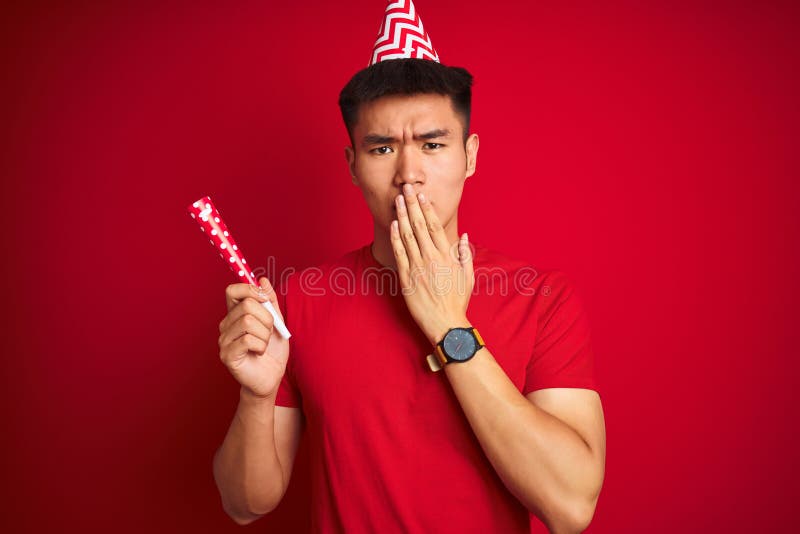 Asian chinese man on birthday celebration wearing funny hat over isolated red background cover mouth with hand shocked with shame