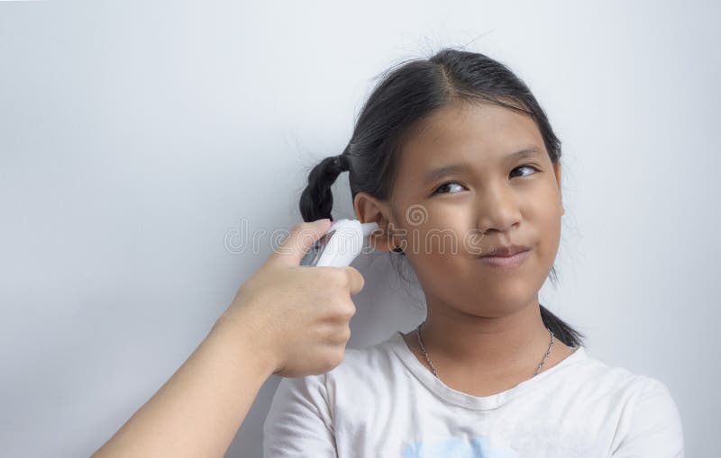 Asian dark-skinned girl smiling awkwardly while doctor measuring her body temperature with digital ear infrared thermometer at clinic