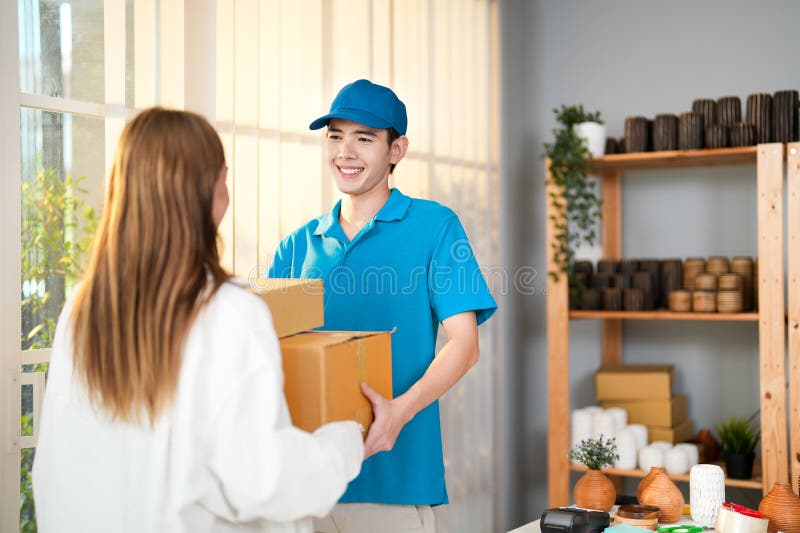 Asian delivery young man in blue uniform giving parcel boxes to business owner woman in her home with vase craftwork background