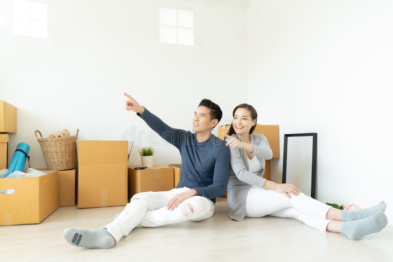 Asian family couple sitting on floor in new apartment with moving boxes, pointing up into the air and smiling with dreaming about stock photos Asian family couple sitting on floor in new apartment with moving boxes, pointing up into the air and smiling with dreaming about stock photos