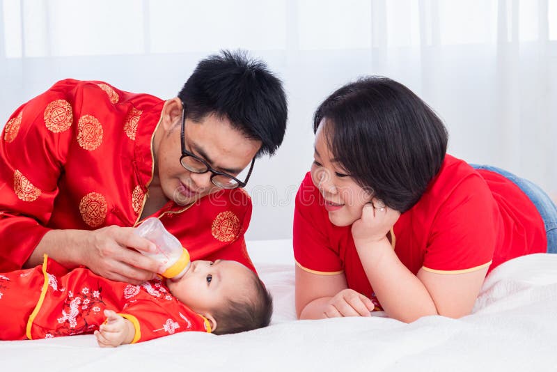 Asian Father Hold Bottle Milk for Son Toddler Bed at Home while Mother Look at Baby with Love New Family Red Chinese Costume Stock Photo
