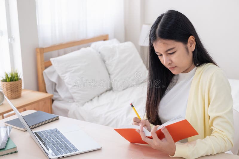 Asian female freelancer reading and jot down note on book while working with laptop on table in bedroom at home.Work at home