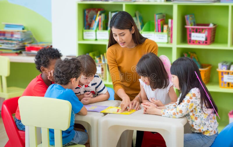 Asian female teacher teaching mixed race kids reading book in cl