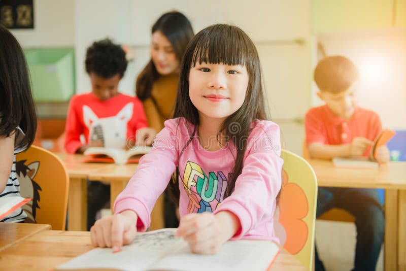 Asian girl reading a book smiling at the camera. Row of multiethnic elementary students reading book in classroom at school. royalty free stock photography