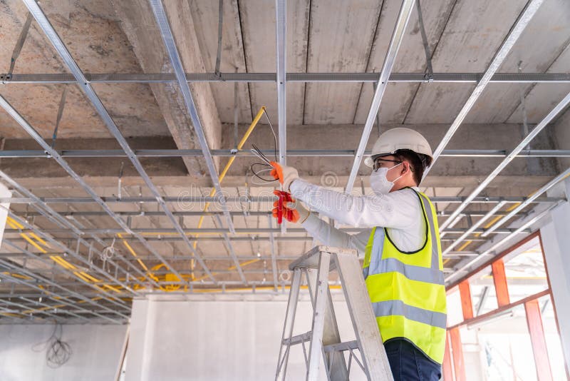 Asian handsome electrician wearing a mask installing laying electrical cables on the ceiling with pliers inside the house under