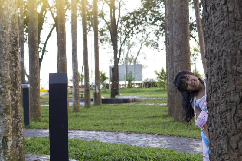 Asian little girl emerges and smile from behind a tree while she playing hide and seek in the park in the evening.