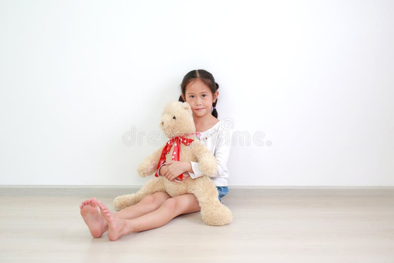 Asian little kid girl hugging a teddy bear doll sitting against white wall in the room with looking at camera