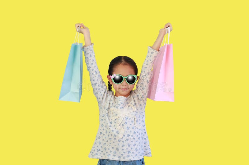 Asian little kid girl wearing sunglasses holding shopping bags isolated on yellow background