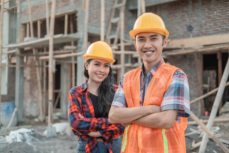 Asian male and female contractors standing with crossed hands smiling at the camera wearing safety helmets