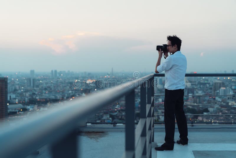 Asian man taking cityscape photo on building rooftop in low light situation. Photography, office people, or hobby concept stock image