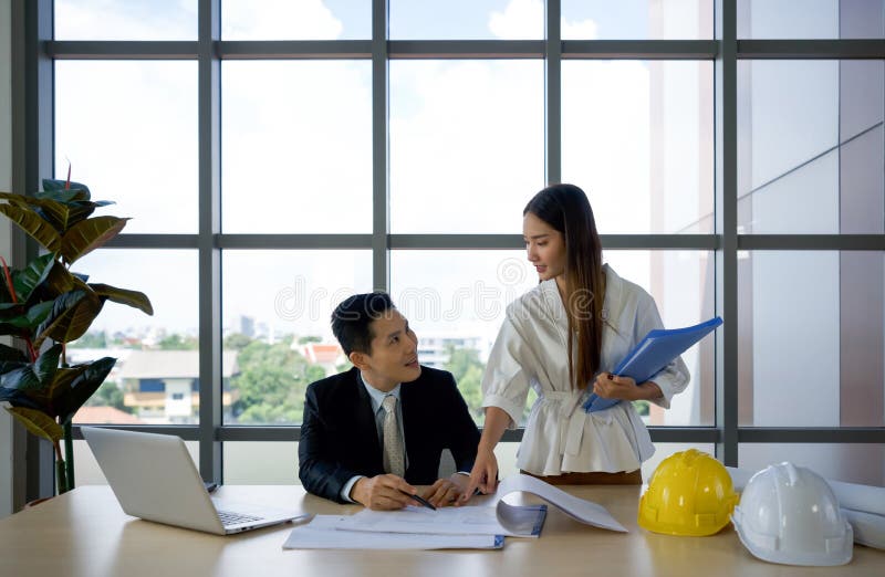 Asian project owner in suit and his assistant checking the construction drawing. Morning work atmosphere in a modern office stock photography