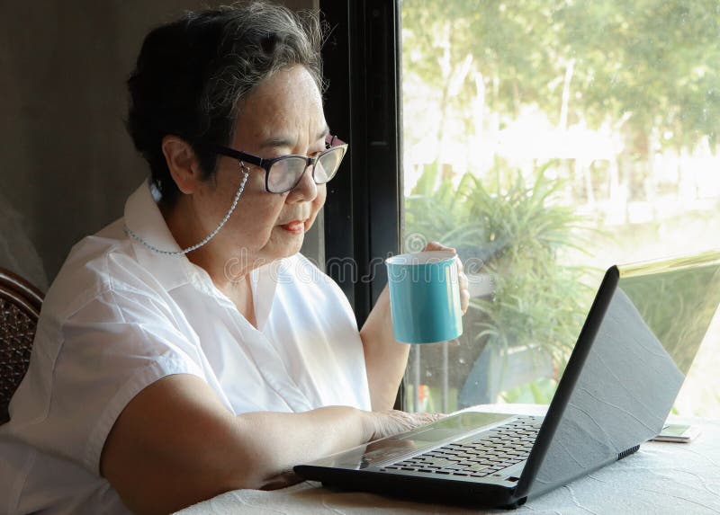 Asian senior woman wearing eye glasses  sitting by window holding blue cup of coffee reading message on computer laptop on  table and smiling. Senior people and technology concept