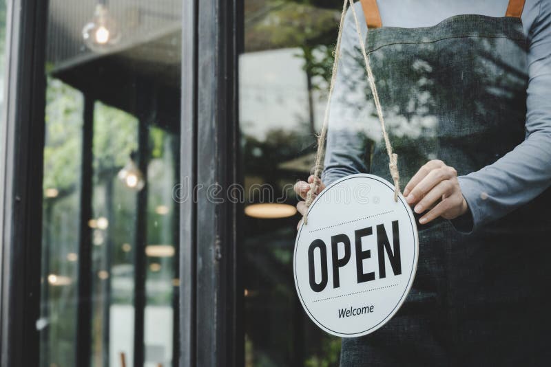 Asian waitress staff woman wearing apron turning open sign board on glass door in modern cafe coffee shop, hotel, cafe restaurant